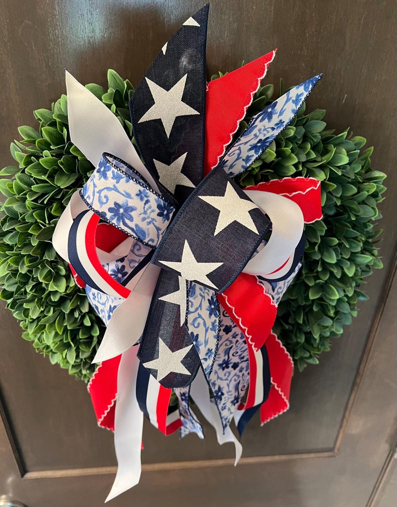 Op de afbeelding: Een decoratieve krans met een dichte groene basis, versierd met patriottische linten. De linten tonen wit, marineblauw met witte sterren, rood en blauwe bloempatronen. Geschikt voor deur- of wanddecoratie.