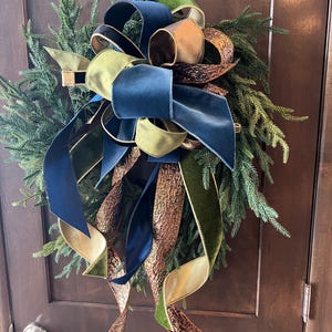 May include: A decorative wreath made of green evergreen branches adorned with a large multi-layered bow. The bow features velvet ribbons in shades of navy blue, olive green, and metallic bronze with a textured, crinkled finish on the long tails.
