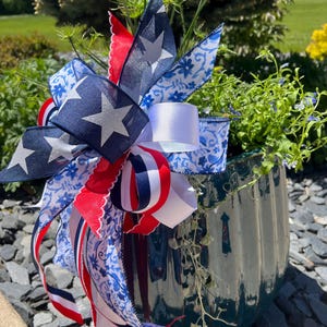 Op de afbeelding: Een decoratieve plantenbak met een patriottisch thema. De donkergroene keramische pot is versierd met een grote strik met marineblauwe linten met witte sterren, rood-wit gestreepte linten en witte linten met blauwe bloempatronen. De plantenbak bevat groen.