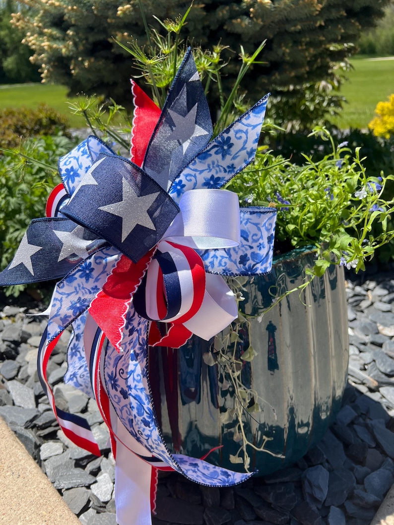 Op de afbeelding: Een decoratieve plantenbak met een patriottisch thema. De donkergroene keramische pot is versierd met een grote strik met marineblauwe linten met witte sterren, rood-wit gestreepte linten en witte linten met blauwe bloempatronen. De plantenbak bevat groen.