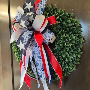Op de afbeelding: Een ronde groene krans versierd met een patriottische strik. De strik heeft linten in rood, wit en blauw, met ster- en bloempatronen. Geschikt voor binnen- en buitengebruik.