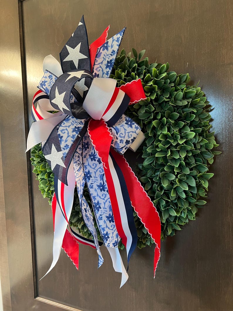 Op de afbeelding: Een ronde groene krans versierd met een patriottische strik. De strik heeft linten in rood, wit en blauw, met ster- en bloempatronen. Geschikt voor binnen- en buitengebruik.