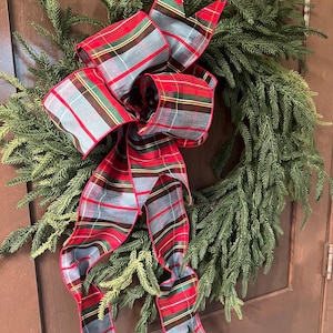 May include: A circular wreath made of dark green evergreen foliage, featuring a large, multi-looped decorative bow. The bow is crafted from fabric with a classic plaid pattern in shades of red, light blue, yellow, and dark green, with long, flowing tails.