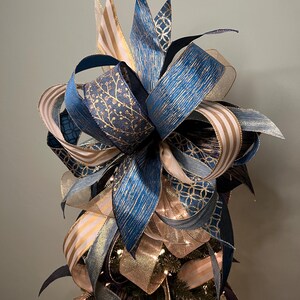May include: A decorative Christmas tree topper featuring layered ribbons in shades of blue, gold, and cream. The ribbons have various patterns, including stripes, floral designs, and geometric shapes. The topper is designed to sit atop a Christmas tree.