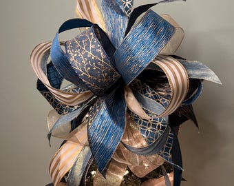 Puede incluir: Un adorno decorativo para árbol de Navidad con cintas en capas en tonos azul, dorado y crema. Las cintas tienen varios patrones, incluyendo rayas, diseños florales y formas geométricas. El adorno está diseñado para colocarse encima de un árbol de Navidad.