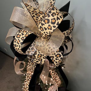 May include: A decorative tree topper featuring a mix of ribbons in leopard print, black, and gold. The leopard print ribbon has a tan background with brown spots. The gold ribbon is sheer. The topper is designed to sit atop a Christmas tree.