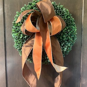 May include: A circular wreath made of dense, dark green boxwood leaves. A large, multi-layered decorative bow is attached to the top, featuring wide ribbons in alternating shades of dark brown and burnt orange with a smooth, satin-like finish.