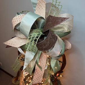 May include: A decorative tree topper featuring layered ribbons in shades of green, brown, and gold. The ribbons are arranged in a bow shape, with some featuring a polka dot pattern and others a textured design. The topper is adorned with small lights.