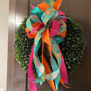 May include: A decorative wreath with a vibrant bow. The wreath features a base of green foliage, adorned with a large bow made of ribbons in shades of orange, teal, pink, and floral patterns. The wreath is designed for door or wall decor.