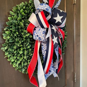 Op de afbeelding: Een ronde, groene buxuskrans versierd met een patriottische strik. De strik heeft linten in rood, wit en blauw, met ster- en bloempatronen. De krans hangt aan een bruine deur en zorgt voor een feestelijke uitstraling.