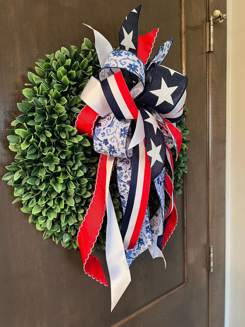 Op de afbeelding: Een ronde, groene buxuskrans versierd met een patriottische strik. De strik heeft linten in rood, wit en blauw, met ster- en bloempatronen. De krans hangt aan een bruine deur en zorgt voor een feestelijke uitstraling.