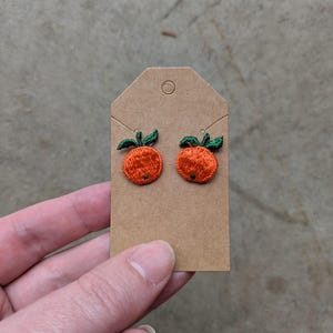 May include: A pair of orange and green embroidered earrings shaped like oranges. The earrings are displayed on a brown card.