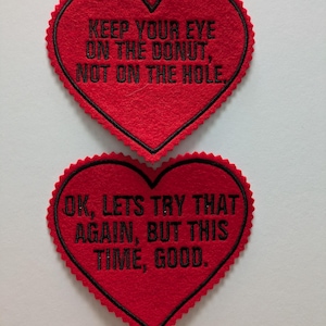 May include: Two red felt heart-shaped patches with black embroidered text. The top patch reads "Keep your eye on the donut, not on the hole." The bottom patch reads "OK, let's try that again, but this time, good."