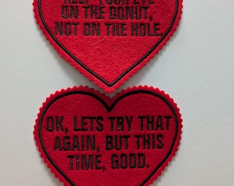 Puede incluir: Dos parches de fieltro rojo en forma de corazón con texto bordado en negro. El parche superior dice "Keep your eye on the donut, not on the hole." El parche inferior dice "OK, let's try that again, but this time, good."