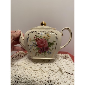 May include: A cream-coloured ceramic teapot featuring a central floral bouquet with pink roses, purple and yellow flowers, and green leaves. The teapot has a gold-trimmed handle, spout, and rim, with a gold-coloured knob on the lid.