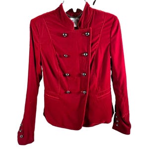May include: A bright red velvet jacket featuring a high collar, long sleeves, and a double-breasted front closure with eight silver-toned metallic buttons. The cuffs of the sleeves also feature three matching silver-toned buttons on each side.