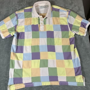 May include: A short-sleeve polo shirt featuring a multi-colored checkerboard pattern in pastel shades of yellow, light blue, green, and purple. The shirt has a solid white collar, a three-button placket, and an Orvis brand label at the neckline.