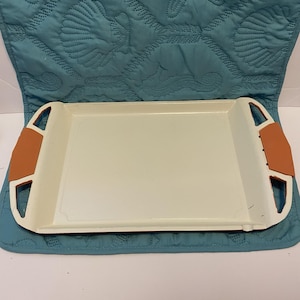 Puede incluir: Una bandeja rectangular de color blanco roto con asas naranjas. La bandeja tiene un borde ligeramente elevado y está colocada sobre una superficie acolchada azul. Las asas son de un color contrastante.