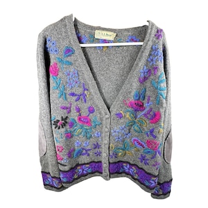 May include: A grey V-neck cardigan sweater featuring colorful floral embroidery in shades of pink, purple, and blue. The garment has a button-front closure, long sleeves with grey elbow patches, and a purple floral band along the bottom hem.