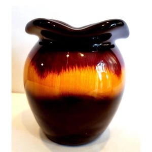 Könnte beinhalten: Eine glänzende Keramikvase mit einem einzigartigen, gerüschten Rand. Die Vase geht von einem tiefbraunen Sockel in einen leuchtend orangefarbenen Mittelteil über und gipfelt in einem dunkelbraunen Oberteil. Das Design ist eine Mischung aus klassischen und modernen Stilen.