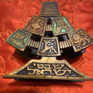 Puede incluir: Objeto decorativo de bronce de varios niveles con una base triangular y paneles cuadrados. Cada panel presenta un diseño en relieve diferente, incluyendo un barco, un león y un amanecer. La base tiene texto hebreo.
