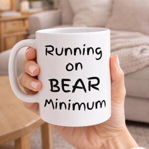 Puede incluir: Taza de cerámica blanca con el texto "Running on BEAR Minimum" en negro. La taza tiene un asa grande y la sostiene una persona. El fondo incluye un sofá y una mesa de madera.