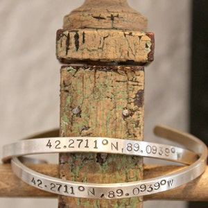 May include: Two silver bracelets with engraved coordinates, "42.2711° N, 89.0939° W", on a weathered wooden surface.