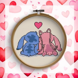 Puede incluir: Un bordado de punto de cruz en un aro de madera presenta una pareja de dibujos animados azul y rosa con un corazón rosa encima. El fondo es blanco con corazones rosas y rojos. El bordado es una artesanía hecha a mano.