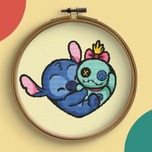 Puede incluir: Un personaje azul Stitch de la película de Disney Lilo & Stitch está abrazando a un personaje Angel verde y azul. Ambos personajes están bordados en una tela blanca en un aro de bordado de madera redondo.