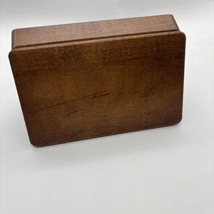 May include: A rectangular wooden box with a smooth, medium-brown finish and visible wood grain. The box features a lid with rounded corners and a clean, simple design, showcasing the natural texture and warm tones of the polished wood surface.