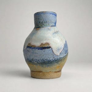 May include: A ceramic vase with a bulbous body and a narrow neck. The surface features a layered glaze with shades of blue, white, and earthy tan. The texture appears mottled and reactive, with a distinct transition between the colors and a visible raw clay base at the bottom.