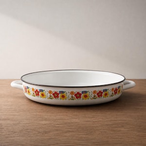 May include: White enamel serving dish with a floral border in red, yellow, and blue. The dish has a black rim and two white handles. The dish is oval-shaped and sits on a wooden surface.