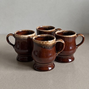 May include: A set of four ceramic mugs with a glossy dark brown finish and a distinctive cream-colored drip glaze around the upper rim. Each mug features a rounded body, a flared base, and a curved handle extending from the side of the vessel.