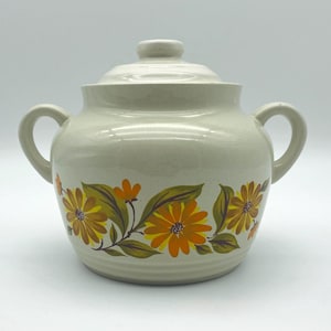 May include: A white ceramic crock pot with a lid, featuring a floral design of orange and yellow flowers with green leaves.