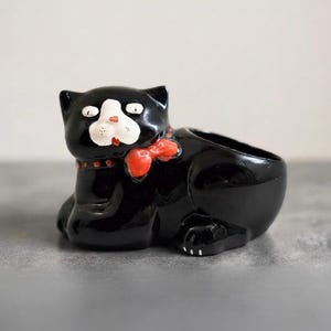 May include: A glossy black ceramic cat planter featuring a white muzzle, small white eyes, and a bright red bow tie around its neck. The cat is depicted in a reclining position with a hollow opening on its back for holding small items or plants.
