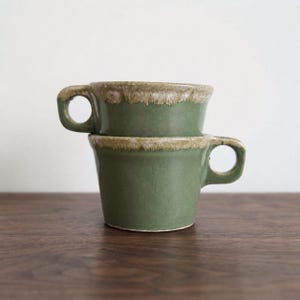 May include: Two stacked ceramic mugs with a sage green glaze and a textured, light beige drip pattern along the upper rim. Each mug features a sturdy, circular handle extending from the side, with a smooth finish on the lower body.