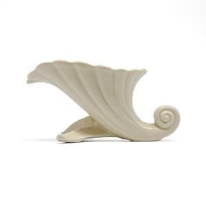 May include: A cream-colored ceramic cornucopia vessel featuring a fluted, shell-like opening and a curled tail end. The piece is supported by a base with two distinct, curved feet that elevate the main body of the horn-shaped decorative object.