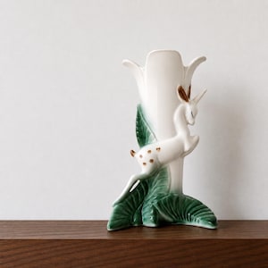 May include: A white ceramic vase with a deer and leaf design. The vase has a tall, tulip-shaped opening and is decorated with a leaping deer with brown spots and antlers. Green leaves accent the base.