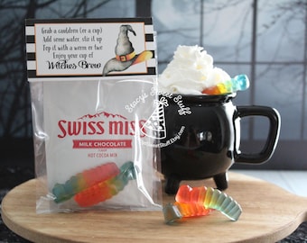May include: A Halloween-themed image featuring a black cauldron mug filled with whipped cream and gummy worms. A packet of Swiss Miss hot cocoa mix and a sign that reads "Witches Brew" are also present. The items are arranged on a wooden serving board.