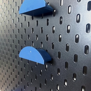 May include: A black pegboard with oval holes. Two blue, semi-circular shelves are attached. The shelves are positioned one above the other, creating a storage or display system. The pegboard is likely for organizing items.