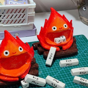 Puede incluir: Figuras hechas a mano de cer&aacute;mica roja y naranja con forma de fuego, con ojos y bocas blancas, sentadas sobre troncos de madera. Incluye peque&ntilde;os bloques blancos con meses y n&uacute;meros. Decorativo y caprichoso.