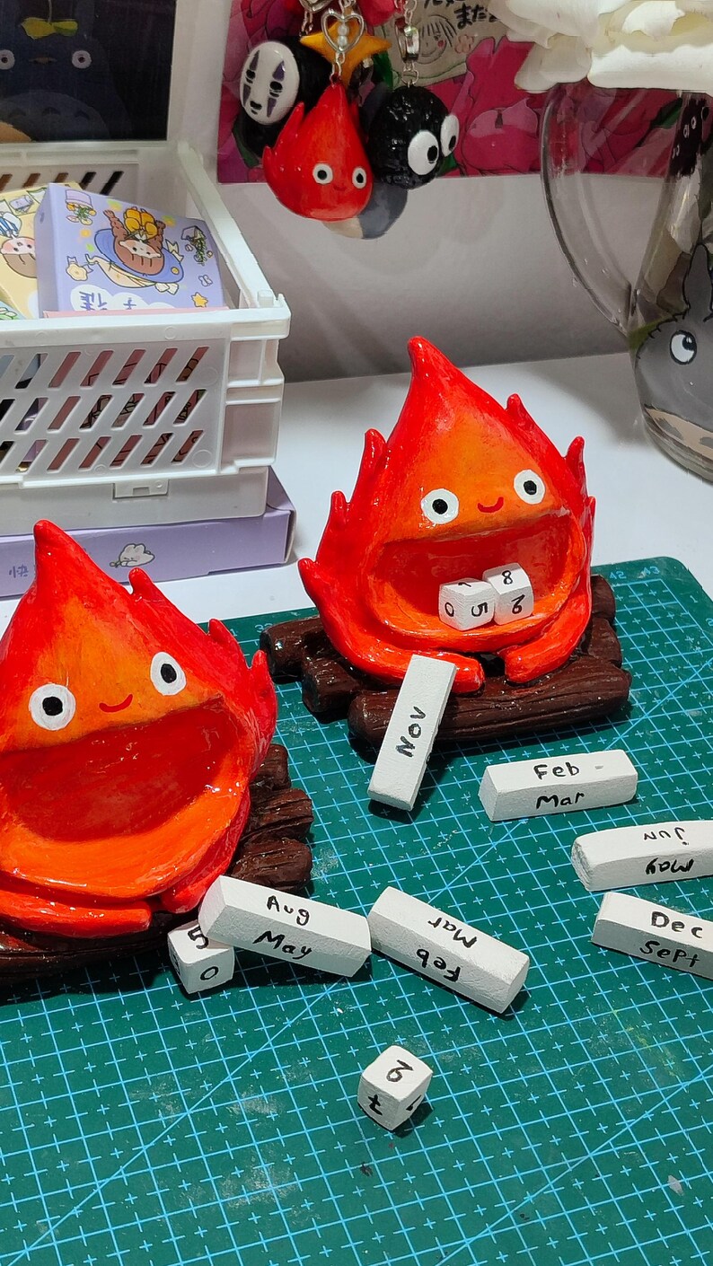 Puede incluir: Figuras hechas a mano de cer&aacute;mica roja y naranja con forma de fuego, con ojos y bocas blancas, sentadas sobre troncos de madera. Incluye peque&ntilde;os bloques blancos con meses y n&uacute;meros. Decorativo y caprichoso.