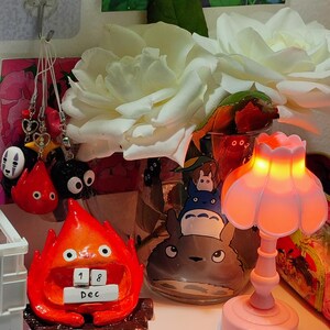 Puede incluir: Una composici&oacute;n decorativa con una figura roja de Calcifer con un calendario que dice "18 Dic", una l&aacute;mpara rosa y un tarro de cristal con personajes de Totoro. Rosas blancas y varios colgantes completan la escena.