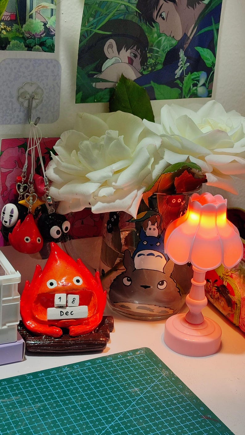Puede incluir: Una composici&oacute;n decorativa con una figura roja de Calcifer con un calendario que dice "18 Dic", una l&aacute;mpara rosa y un tarro de cristal con personajes de Totoro. Rosas blancas y varios colgantes completan la escena.