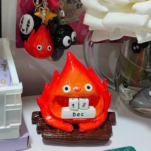 Puede incluir: Una figura artesanal de Calcifer rojo y naranja de El castillo ambulante, con la fecha del 18 de diciembre. Se ven amuletos adicionales y una rosa blanca en el fondo.