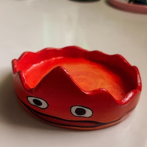 May include: A red and orange ceramic dish with a wavy rim and two black eyes painted on the front. The dish is shaped like a crown.