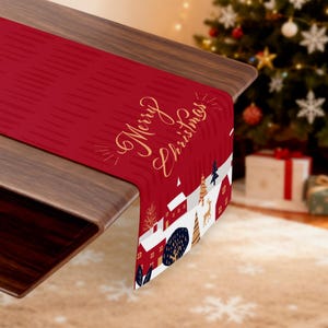 Może przedstawiać: Czerwony bieżnik świąteczny z napisem "Merry Christmas" w złotym kolorze. Bieżnik przedstawia zimową scenę wioski z domami, drzewami i jeleniami. Umieszczony na drewnianym stole.