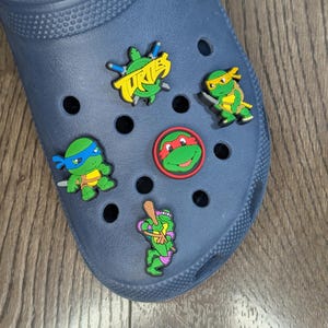 Puede incluir: Zapato azul con cinco dijes de zapatos con temática de las Tortugas Ninja. Los dijes muestran a las tortugas en diferentes poses, incluyendo una con un bate de béisbol, una con una katana y una con un sai. Los dijes también muestran la palabra "Turtles" en letras amarillas.