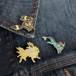 Puede incluir: Tres pines de esmalte con personajes de dibujos animados. Un pin es negro y dorado con un diseño de conejo, otro es amarillo y dorado con un diseño de zorro, y el tercero es azul con un diseño de pez. Los pines están sobre una chaqueta vaquera azul.