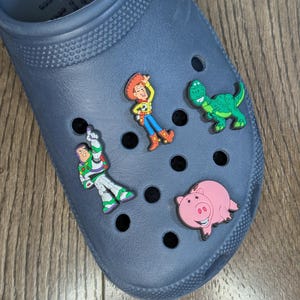 Peut inclure: Un sabot bleu marine orné de breloques de personnages colorés. Les breloques comprennent Buzz l'Éclair, Woody, un dinosaure vert et un cochon rose. La chaussure est posée sur un fond en bois.